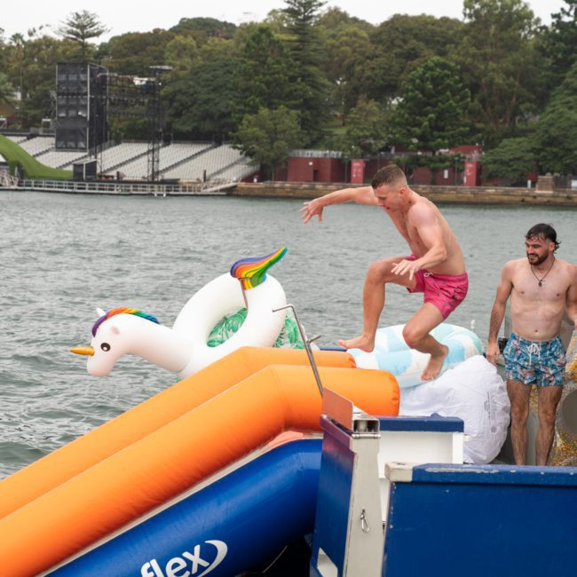 A man in pink shorts jumps into the water from a boat, aiming for an inflatable unicorn, while three others watch. The background shows a shoreline with trees and structures, capturing the fun of DJ boat hire Sydney events.