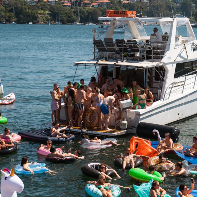 A group of people are gathered on and around a docked boat, floating on various inflatable devices in the water. With greenery and houses visible in the background, it's an ideal setting for luxury yacht hire Sydney events.
