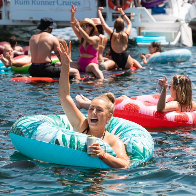 People on colorful inflatable floats enjoying a sunny day on the water, one person in the foreground raising a hand in a wave while holding a drink. This scene perfectly captures the fun ambiance of Sydney boat party hire, creating unforgettable moments against the vibrant backdrop of summer festivities.