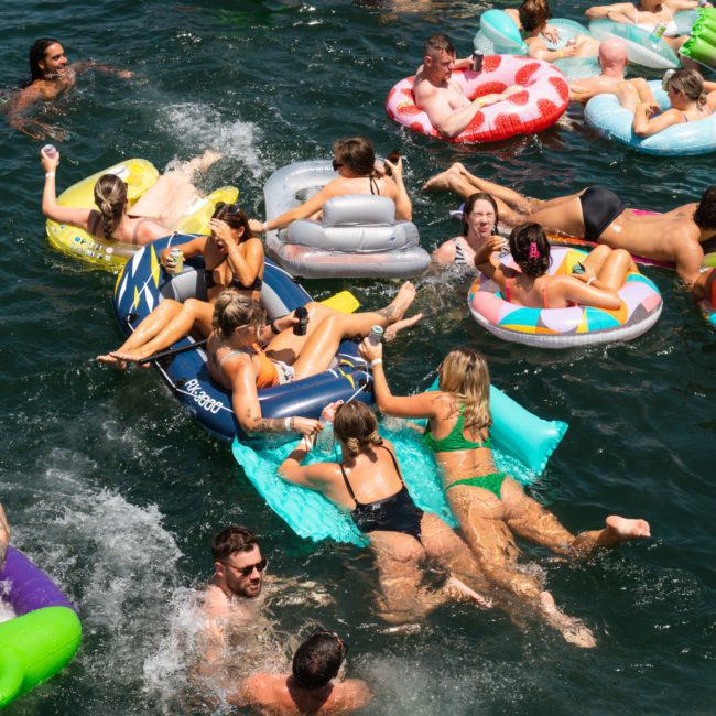 Numerous people are swimming and lounging on colorful floaties in a body of water, enjoying a sunny day near a luxury yacht hire in Sydney.