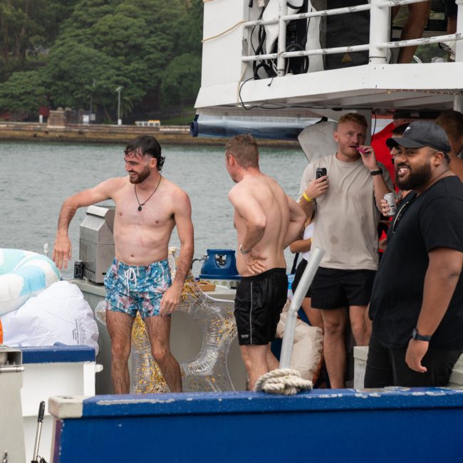 People on a Catamaran party Sydney, some wearing swimsuits, are enjoying a gathering on the water. One man is wet and standing near inflatables. Others are laughing, drinking, and taking photos.