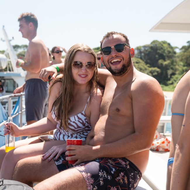 A man and woman in swimwear sit together, smiling and holding drinks on a boat with other people in the background at a lively catamaran party in Sydney.
