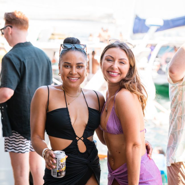 Two women in swimwear pose together on a boat, smiling at the camera. One holds a can of drink. Other people and boats are visible in the background, hinting at a lively Sydney boat party hire event.