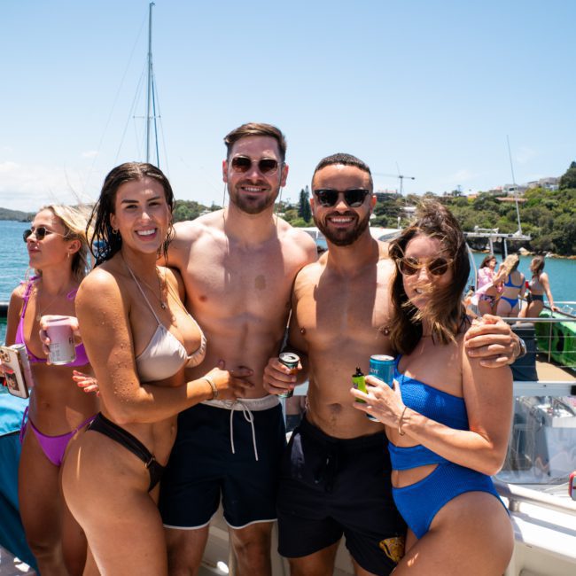 A group of people in swimwear standing and smiling on a boat, holding drinks. The background shows water, more boats, and a partly cloudy sky, capturing the essence of a Sydney boat party hire experience.