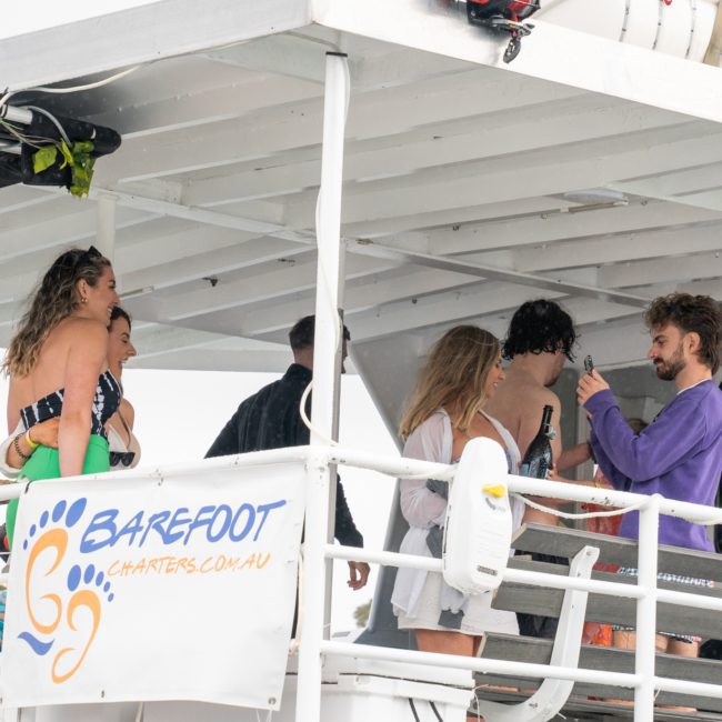 People are gathered on the deck of a boat named "Barefoot Charters." Some are chatting and holding drinks under a shaded area. The luxury yacht hire in Sydney creates a perfect ambiance, with the boat docked and land visible in the background.