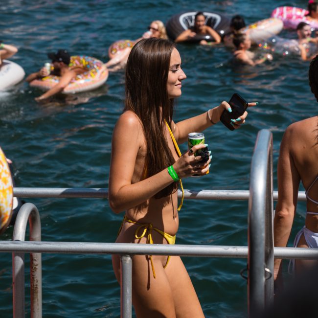 Person in a yellow swimsuit holding a drink stands on a dock near a water area filled with people on colorful floats and a luxury yacht hire Sydney.
