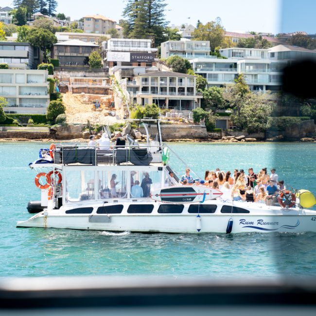 A group of people are seen on a luxury yacht named River Romance, cruising on clear blue water near a coastline with modern buildings and lush greenery in the background. Perfect for those looking for an unforgettable Sydney boat party hire experience.