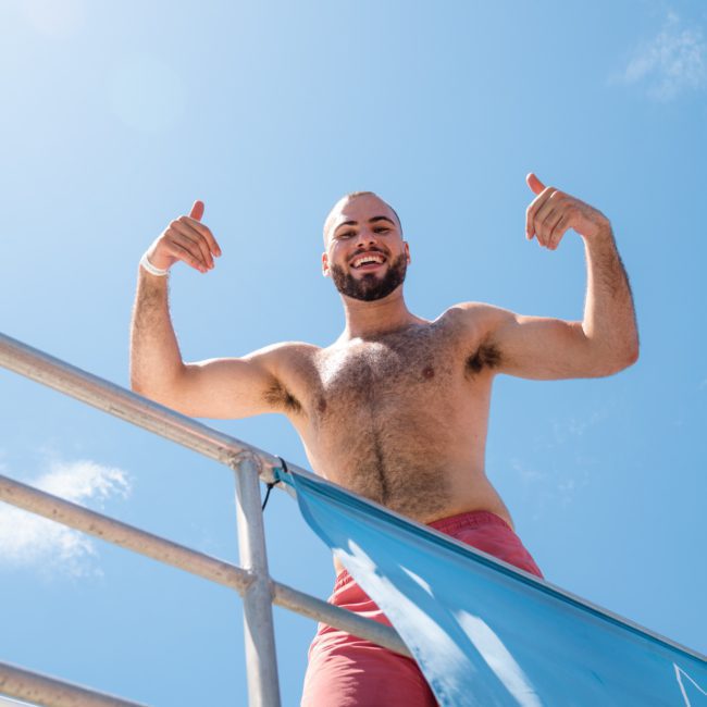 A man in red shorts stands on a metal railing platform against a clear blue sky, raising his hands and smiling at a Sydney boat party hire.