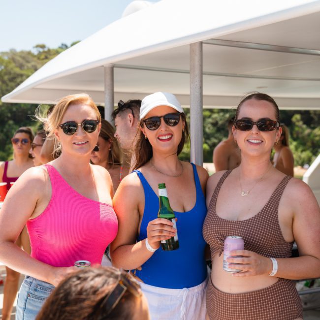 Three women in swimsuits and sunglasses standing on a private yacht charter in Sydney Harbour, holding drinks, with a group of people in the background.
