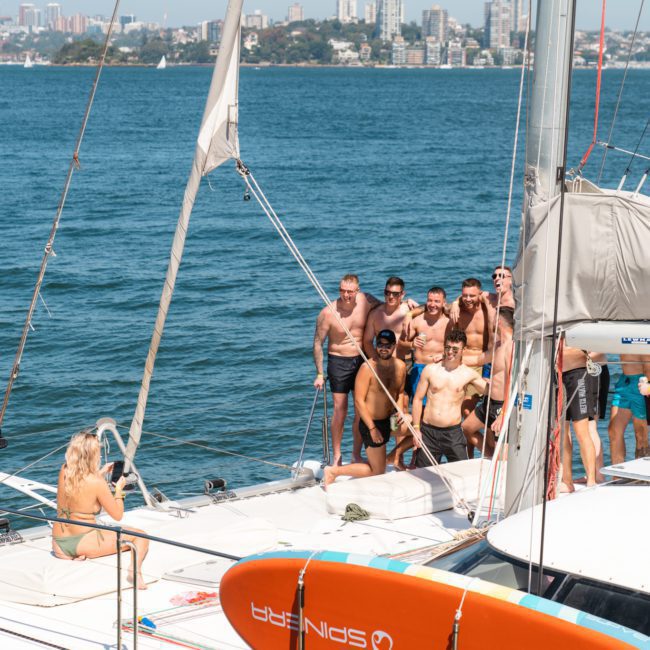 A group of people in swimsuits socializing on a luxury yacht hire Sydney near the coastline, with city buildings visible in the background.