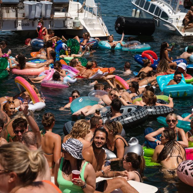 A large group of people are enjoying a summer day in the water, surrounded by colorful inflatable floats, with boats docked nearby. It looks like a festive Sydney boat party hire or perhaps an exciting corporate boat event on Sydney Harbour.