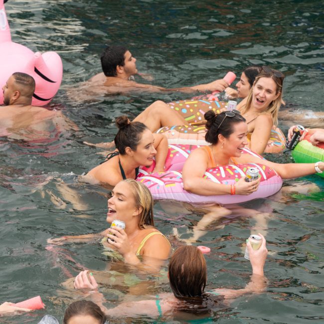 A group of people enjoys a Sydney boat party hire, floating on inflatable rafts and chatting. Various inflatables and drinks are visible, creating an inviting atmosphere on the water.