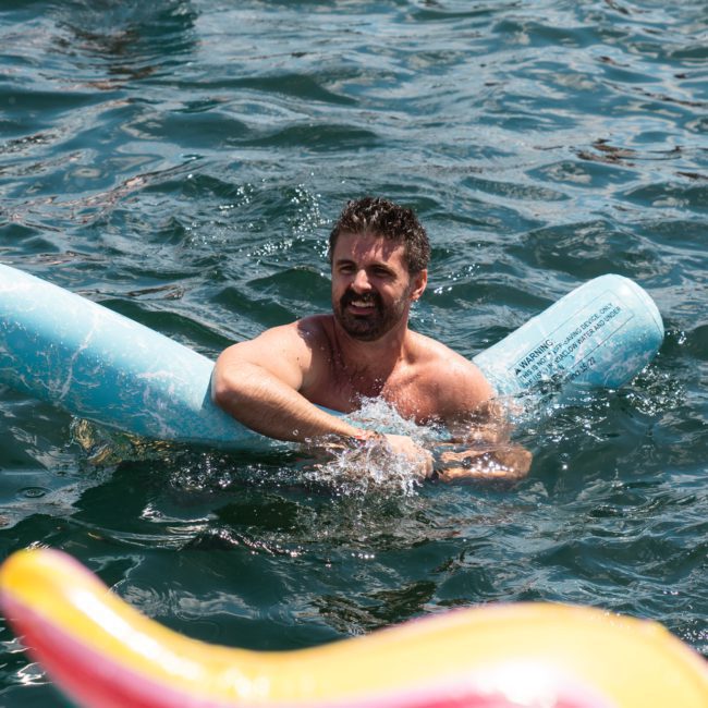 A man swims in the water using a blue pool noodle for support. He appears to be enjoying himself on a sunny day next to a luxury yacht hire in Sydney.