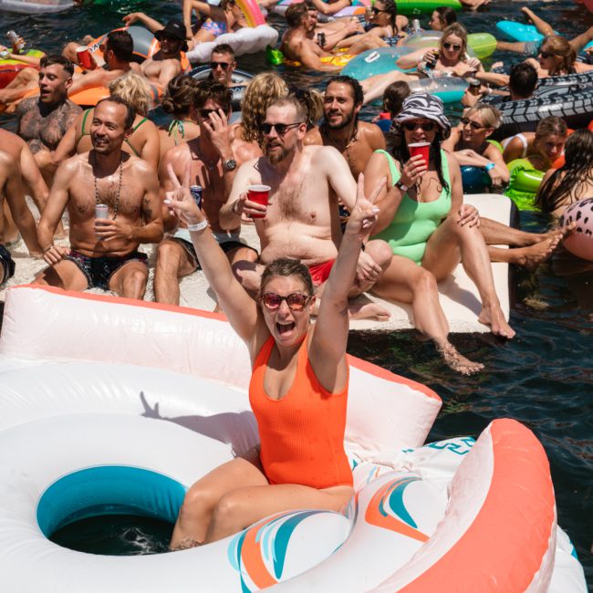 People floating in a lake on inflatable pool floats, some holding drinks. One person in an orange swimsuit in the foreground raises hands, making a peace sign and smiling, reminiscent of the lively atmosphere at corporate boat events Sydney.