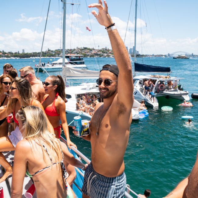 A group of people on a boat are enjoying a sunny day, with some holding drinks. Other boats are visible on the water, along with city buildings and a bridge in the background. It looks like a perfect setting for a private yacht charter Sydney Harbour event.