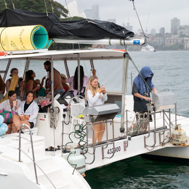 A group of people relax and take photos on a luxury yacht hire Sydney in a harbor on an overcast day. The city skyline is visible in the background.