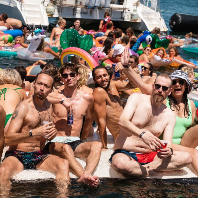 A large group of people in swimsuits are having a catamaran party on a sunny day, surrounded by colorful inflatable floats near boats. They are smiling, raising drinks, and enjoying the water.