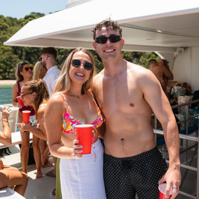 Two people smiling in sunglasses, one wearing a colorful top and white skirt, the other shirtless and wearing black shorts. Both are holding red cups on a catamaran party in Sydney with others in the background.