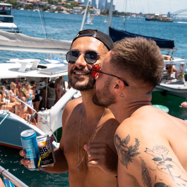 Two men are standing on a boat, one holding a can and the other appearing to lean in close. They are surrounded by other boats and people in swimwear on a sunny day, enjoying a private yacht charter Sydney Harbour with its stunning backdrop.