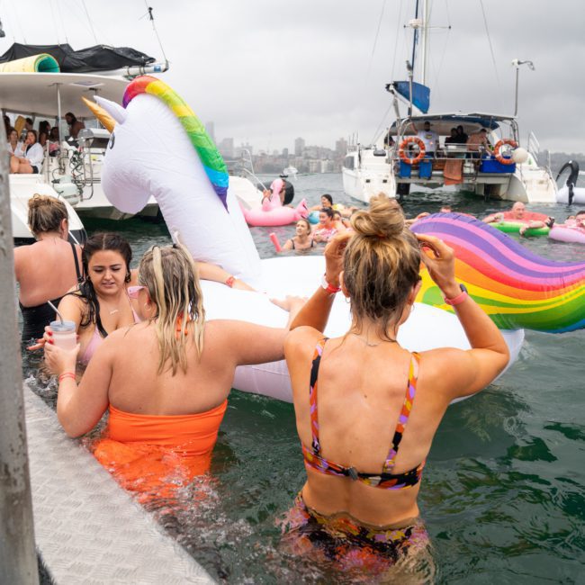 People in swimsuits enjoying the water near boats, with some holding onto a large unicorn inflatable float. The scene appears to be a catamaran party on a cloudy day with a cityscape in the background.