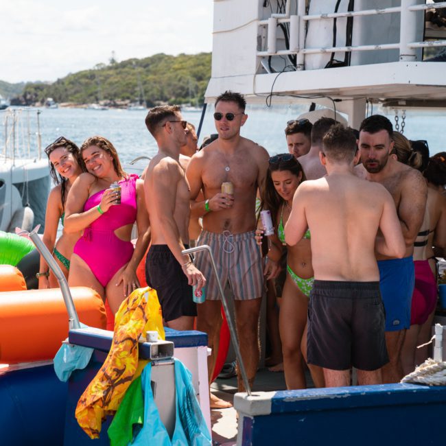 A lively group in swimwear is gathered on a boat, socializing and enjoying drinks. Colorful water inflatables can be seen in the foreground, with the shoreline in the background. It's a perfect scene for a catamaran party Sydney or even considering private yacht charter Sydney Harbour for your next event.