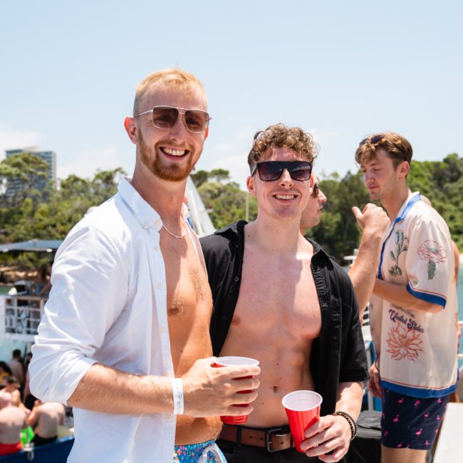 A group of people enjoy a sunny day on a boat. Two men in the foreground, one in a white shirt and the other shirtless with sunglasses, hold red cups. Other people and boats are visible in the background, showcasing what could be an unforgettable Sydney boat party hire experience.