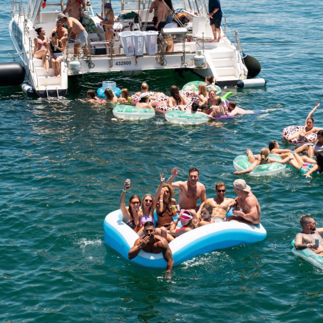People are enjoying a sunny day on the water, swimming, and lounging on inflatables near anchored boats. Some are raising their hands and posing for a photo during a Catamaran Party in Sydney.
