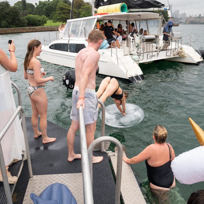 People are enjoying a catamaran party in Sydney. Some stand on the boat platform, one dives into the water, another uses a floaty unicorn, and others are on a nearby boat.