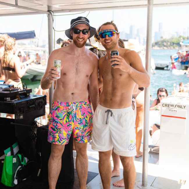 Two men in swimwear, one in colorful shorts and sunglasses, the other in white shorts and visor, pose with drinks on a luxury yacht hire Sydney with a party scene and city skyline in the background.