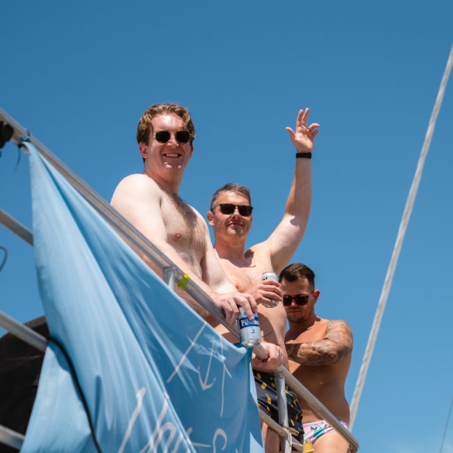 Three men are standing on a boat deck under a clear blue sky. One man waves at the camera, another holds a drink, and the third looks on. A blue banner is draped over the railing in front of them, advertising DJ boat hire Sydney for an unforgettable experience.