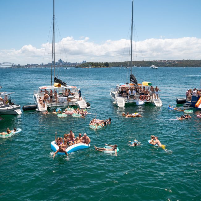 A group of people on inflatables and boats enjoy a sunny day in the water, with a city skyline and bridge in the background, highlighting the perfect setting for a catamaran party on Sydney Harbour.