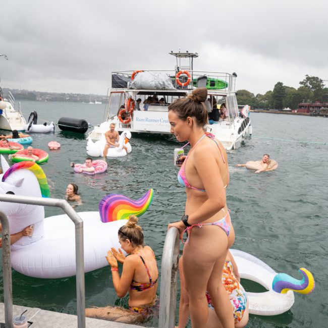 People are enjoying a catamaran party in Sydney on the water. Some are on floats, including a unicorn float, while others are on the boat. The sky is overcast. With Sydney boat party hire services, this event offers a perfect blend of fun and relaxation amidst the scenic views.