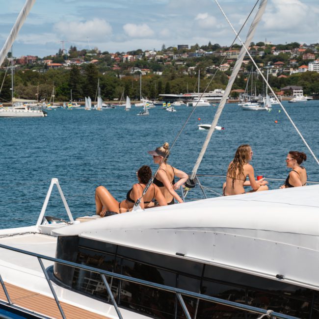 Four people in swimwear relaxing and socializing on the deck of a private yacht charter Sydney Harbour with a scenic view of a bay filled with other boats and a coastline in the background.