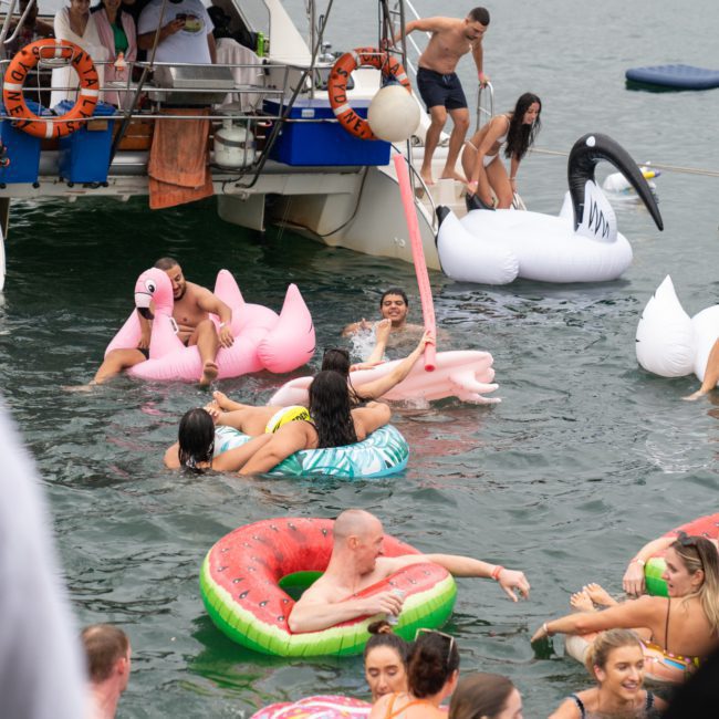 People are enjoying themselves on various inflatable floats in the water near a boat. Some floats are shaped like swans and watermelons, creating a lively and fun atmosphere. Consider enhancing your experience with a private yacht charter in Sydney Harbour for the ultimate celebration.