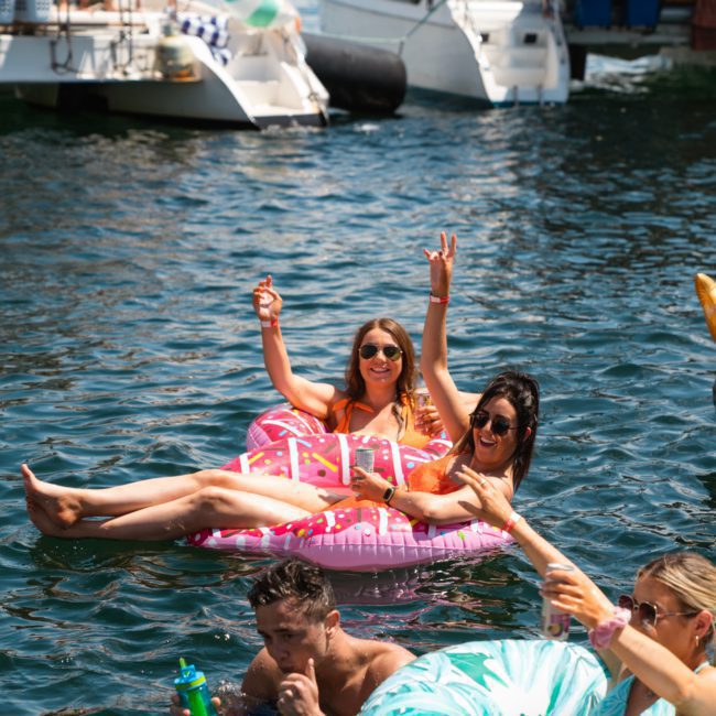 People enjoying a day in the water with inflatable floats, surrounded by boats. Two women float on a donut-shaped raft, raising their hands in celebration during a catamaran party Sydney event while others swim and relax nearby.