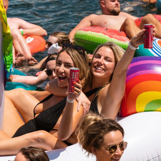 Two people in swimsuits hold drinks while sitting on inflatable pool floats, enjoying a DJ boat hire Sydney experience with others around them in the water.