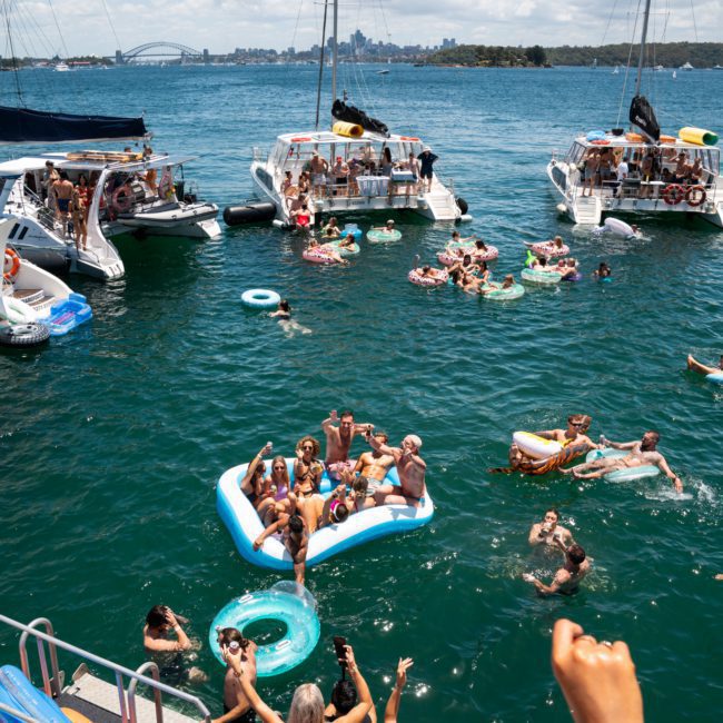 A group of people enjoying a sunny day on an anchored boat, swimming and floating on inflatables in the clear blue waters, with other boats and a city skyline visible in the background. Perfect for a Sydney boat party hire or corporate boat events Sydney, creating unforgettable moments.