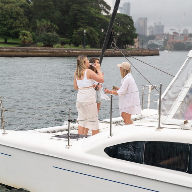 Three people, dressed in light summer outfits, stand on the deck of a white yacht near a forested shoreline with a cityscape in the background on a cloudy day, enjoying a private yacht charter Sydney Harbour.