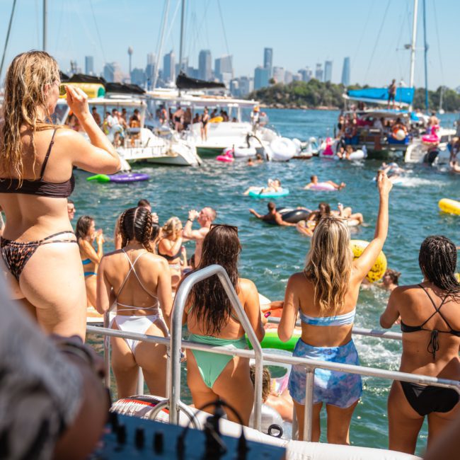 People in swimsuits enjoy a sunny day on a crowded Catamaran party Sydney, with many floating in the water. The city skyline is visible in the background.