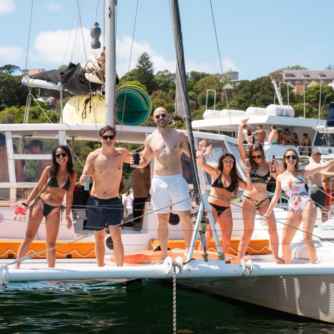 A group of people in swimwear stand and pose on a catamaran during a Private yacht charter Sydney Harbour, docked in a busy marina on a sunny day.