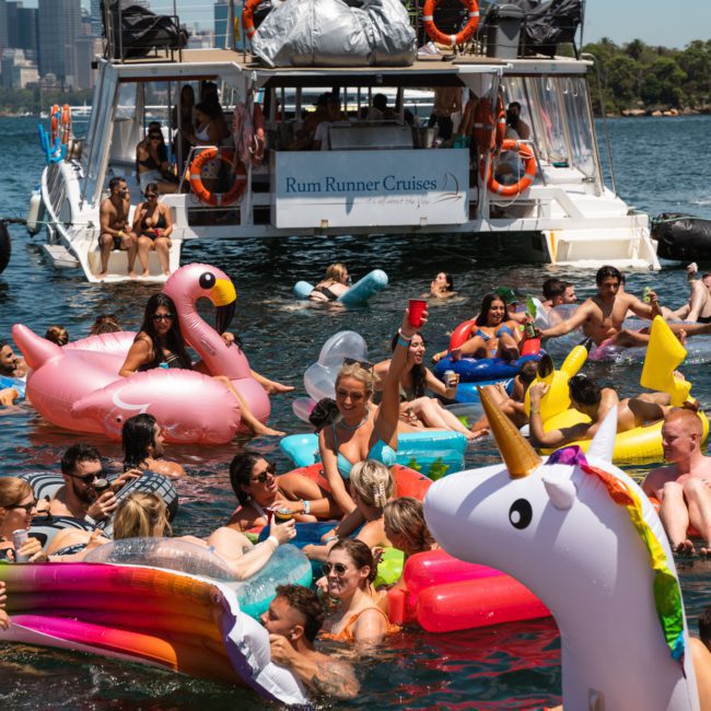 A group of people are enjoying a Sydney boat party hire on the water with inflatable floats, near a boat labeled "Rum Runner Cruises." They are swimming and socializing under a clear sky.