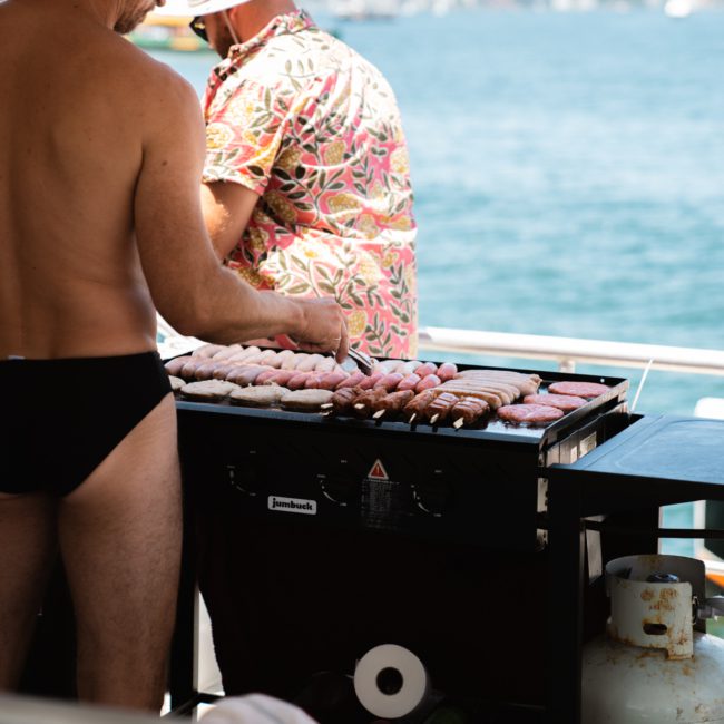 Two people are grilling assorted meats on a barbecue grill on a boat. The person in the foreground is shirtless and wearing swim trunks, while the background shows water and boats, suggesting a vibrant Sydney catamaran party.