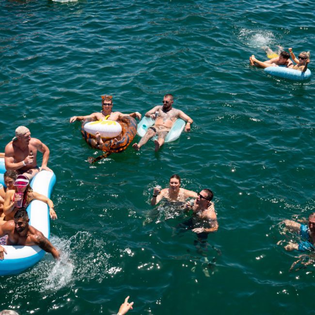 Group of people swimming and floating on inflatables in a body of water on a sunny day near a private yacht charter Sydney Harbour. Some are in a blue inflatable raft and others are on individual floaties, enjoying the serene atmosphere.