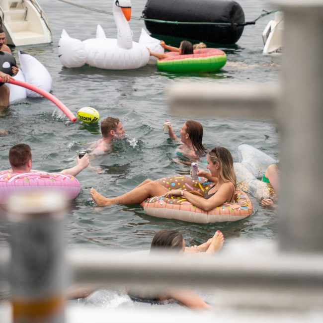 People are floating on various inflatable tubes in a body of water, with one person on a swan floatie and others on donut and pizza-shaped inflatables, enjoying a social gathering. Consider enhancing the experience with DJ boat hire Sydney for an unforgettable party atmosphere.