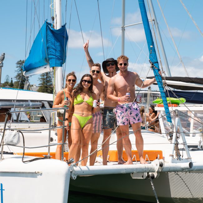 A group of people in swimwear stand on the bow of a sailboat named "Catalyst," smiling and posing for the camera on a sunny day. Enjoying their private yacht charter Sydney Harbour, they epitomize the perfect escape and fun in the sun.