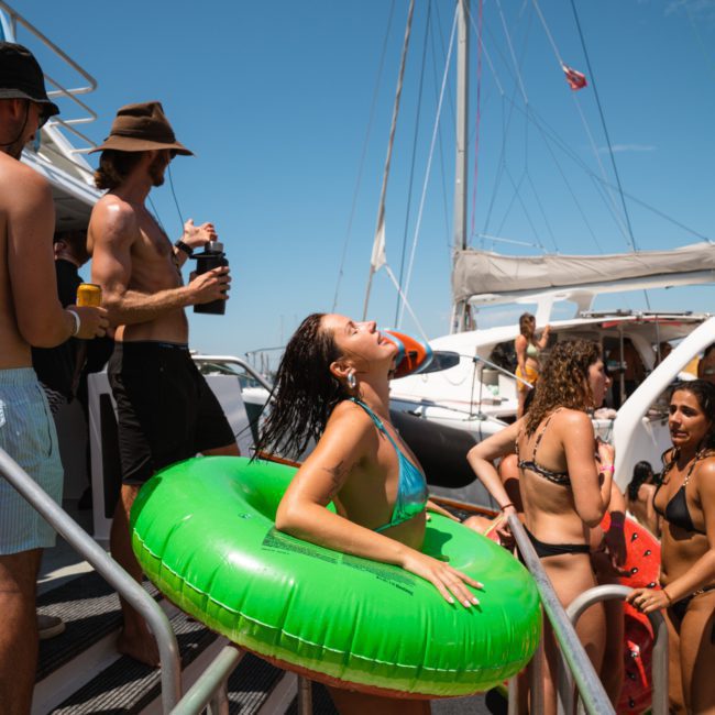 People in swimsuits on a boat deck, enjoying a sunny day. A woman is wearing a green inflatable ring while others stand and socialize around her. Sailboat masts and a bright blue sky are visible—perfect for those considering Luxury yacht hire Sydney to elevate their experience.