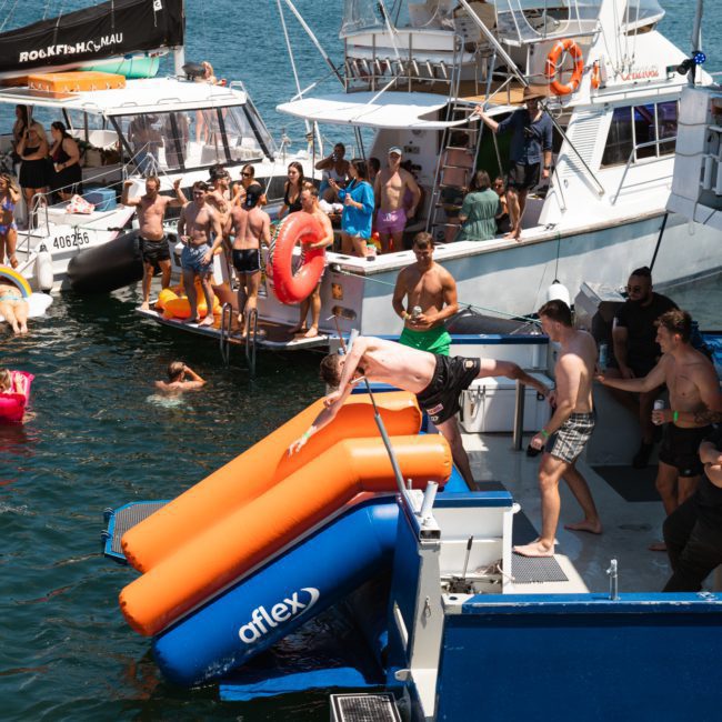 People are enjoying a sunny day on boats and in the water, with some using an inflatable slide to enter the sea. It looks like a perfect Sydney boat party hire, ideal for those wanting to bask in the beauty of Sydney Harbour on a lively adventure.