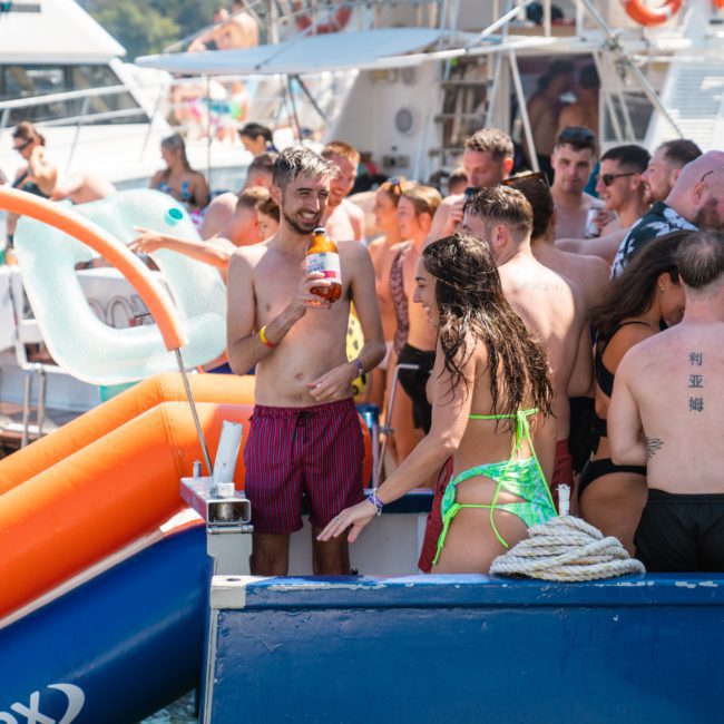 A group of people in swimsuits are gathered on a boat, some with drinks in hand. Several boats are nearby, and an inflatable slide is attached to one of them. The scene appears festive, perfect for a lively Sydney boat party hire or a catamaran party Sydney.