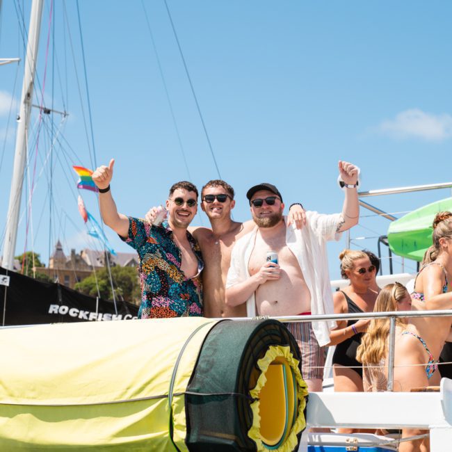 A group of people standing on a boat, with three men in the center giving thumbs up. The boat, suitable for a Sydney boat party hire, is equipped with various gear and features a rainbow flag. The weather is sunny with clear skies.