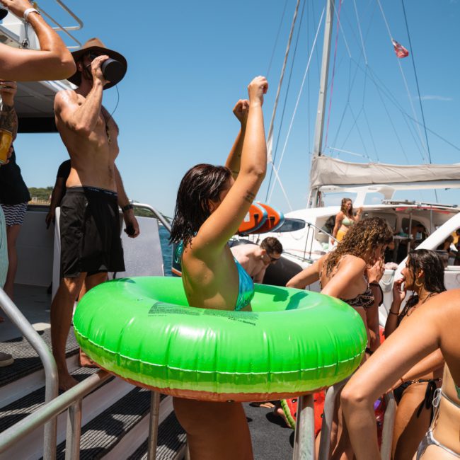 A group of people wearing swimsuits enjoy a sunny day on a boat. One person in the center wears a green inflatable ring. Other boats and clear skies are visible in the background, perfect for those looking into private yacht charter Sydney Harbour or Sydney boat party hire.